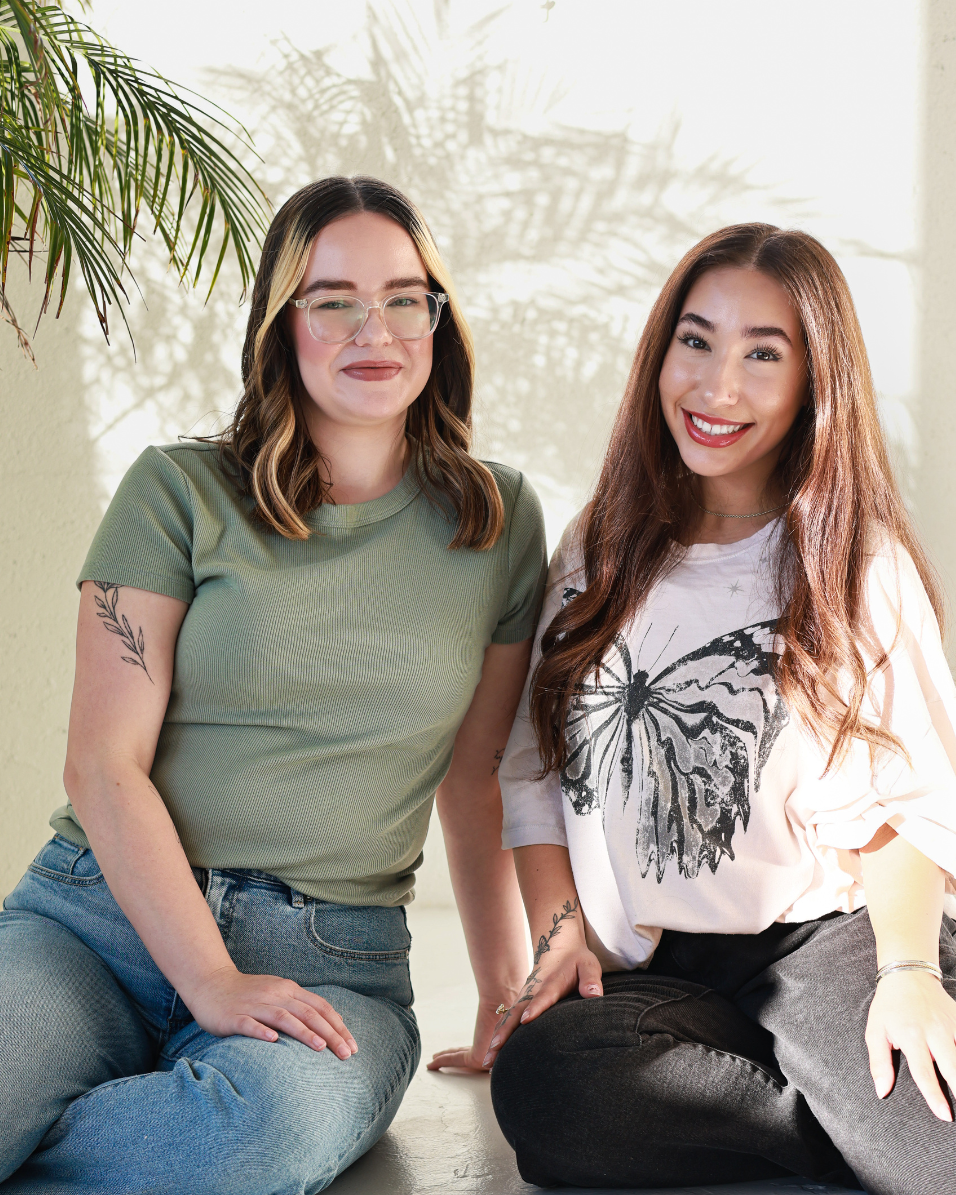 Two women sitting on the floor indoors with a plant in the background